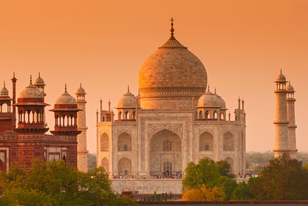 The iconic Taj Mahal, a magnificent white marble mausoleum, stands prominently at the end of a long reflecting pool flanked by manicured green lawns and cypress trees under a clear blue sky.